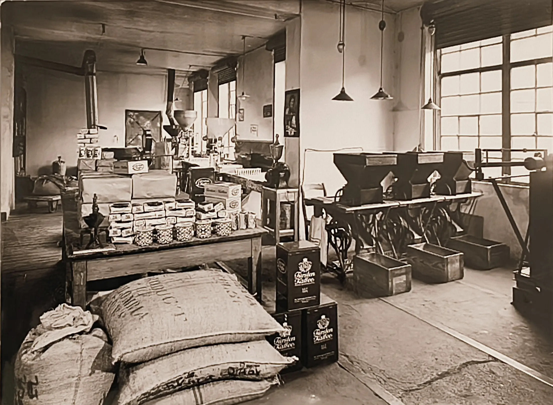 Innenansicht der historischen Rösterei: Kaffeesäcke, Verpackungen und Abfüllmaschinen vor großen Sprossenfenstern