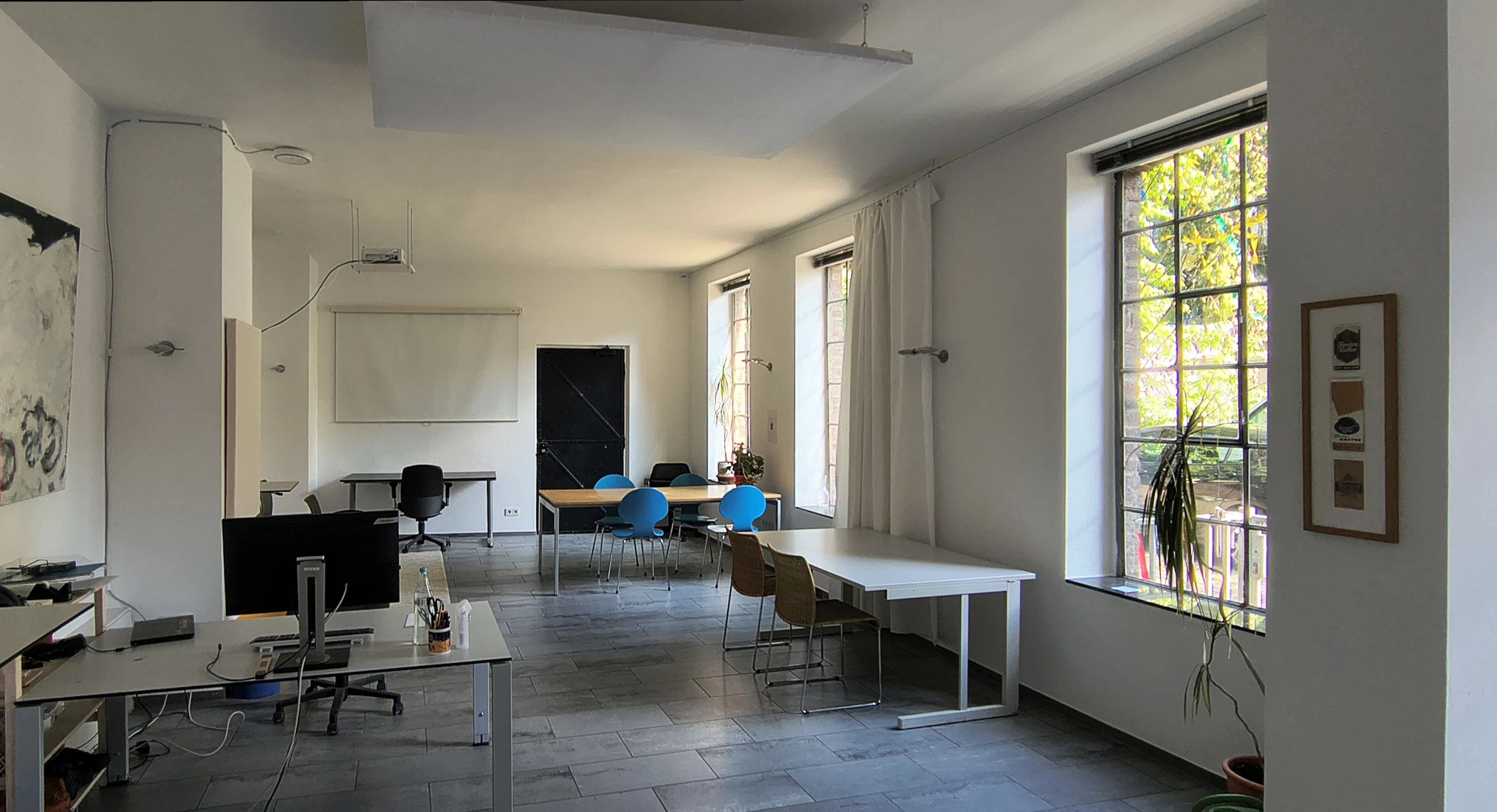 Blick durch den Raum: Schreibtische, Leinwand mit Beamer, Sprossenfenster und schwarze Eisentür